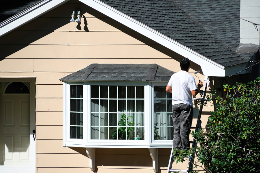 man standing on ladder in front of a bay window. The house has tan siding and a black roof